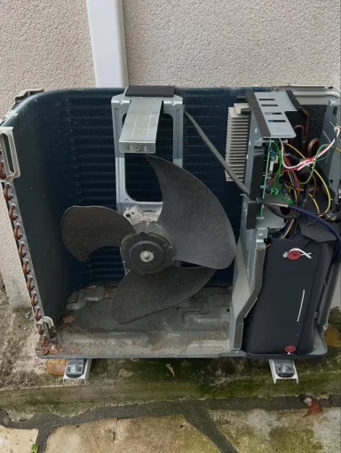 Unité extérieure de climatisation démontée au sol, exposant l'hélice du ventilateur et les composants internes.